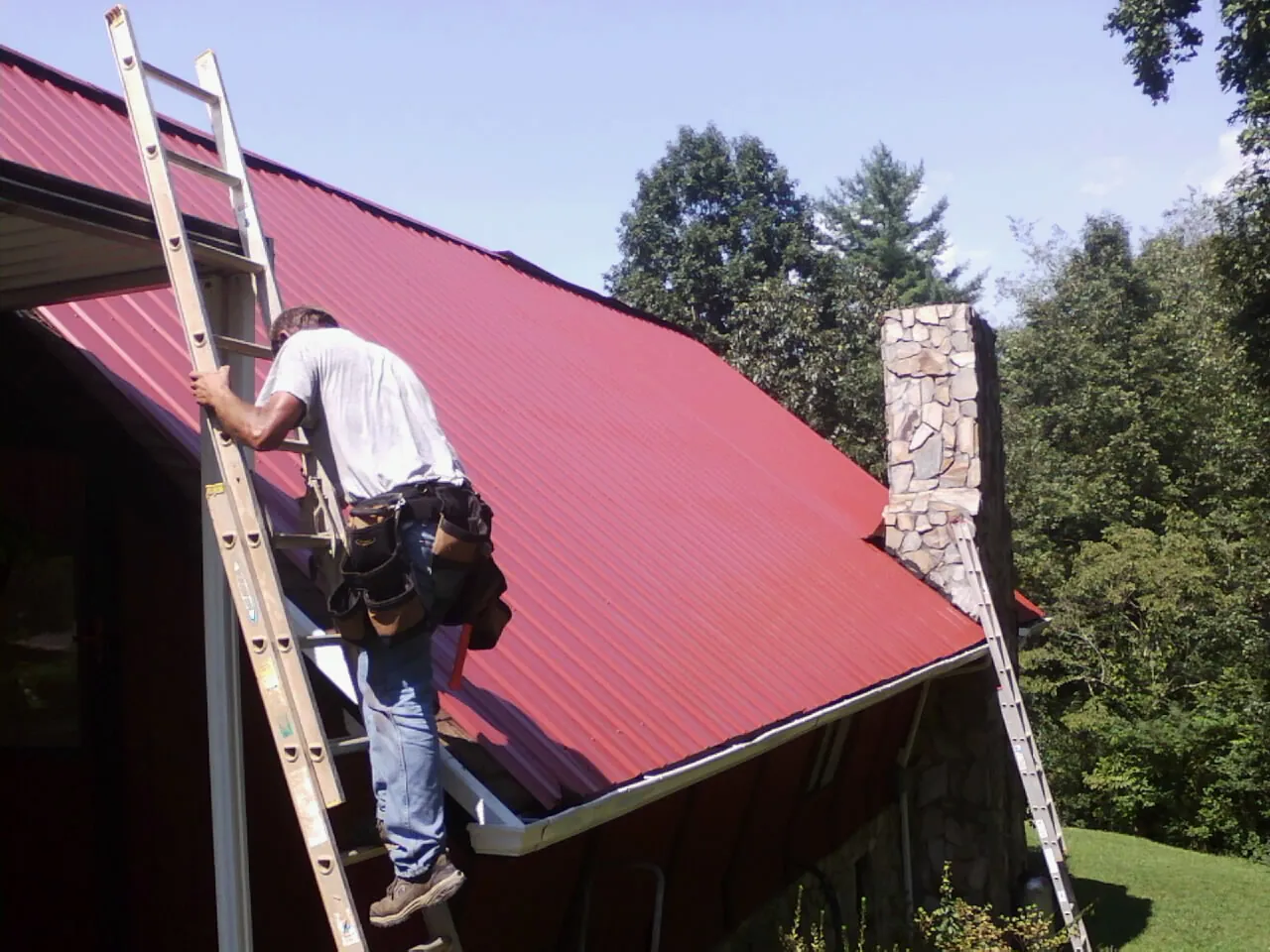 Roofer on a ladder beside a red metal roof and stone chimney — Soffit & Fascia Repair by Roofing Brothers in Averill Park
